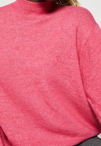 Pull en tricot rose avec un col haut, une texture douce et une coupe décontractée. Côtes visibles au niveau du col et des poignets.