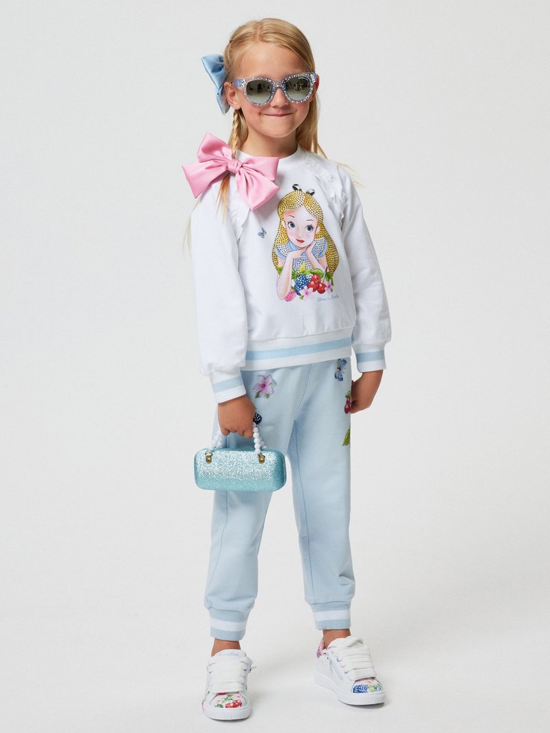 Felpa bianca con stampa di cartoni animati, dettaglio di fiocco rosa, abbinata a pantaloni jogger azzurri chiari con applicazioni floreali e una borsa blu glitterata.