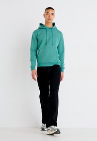 Teal hooded sweatshirt with a front kangaroo pocket, paired with black trousers and multi-colored sneakers. Smooth texture and relaxed fit.