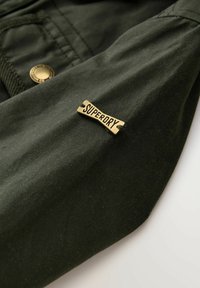 Mörkgrön tyg med en rektangulär mässingsetikett sydd på, präglad med ordet "SUPERDRY" i svarta bokstäver.