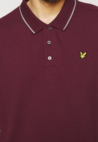 Polo couleur marron avec trois boutons, rayure grise sur le col et petit logo d'oiseau jaune sur le côté gauche de la poitrine.