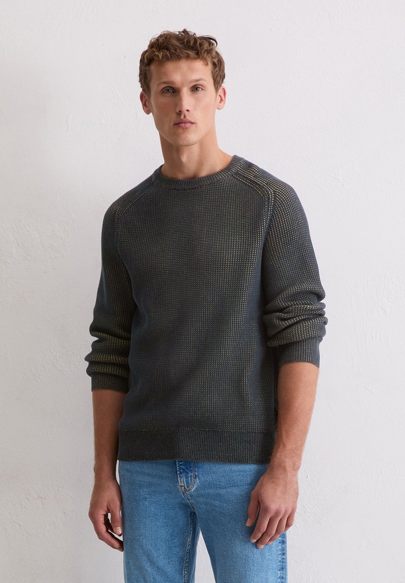 Jeune homme aux cheveux bouclés portant un pull texturé sombre et un jean bleu clair, debout contre un mur blanc uni.