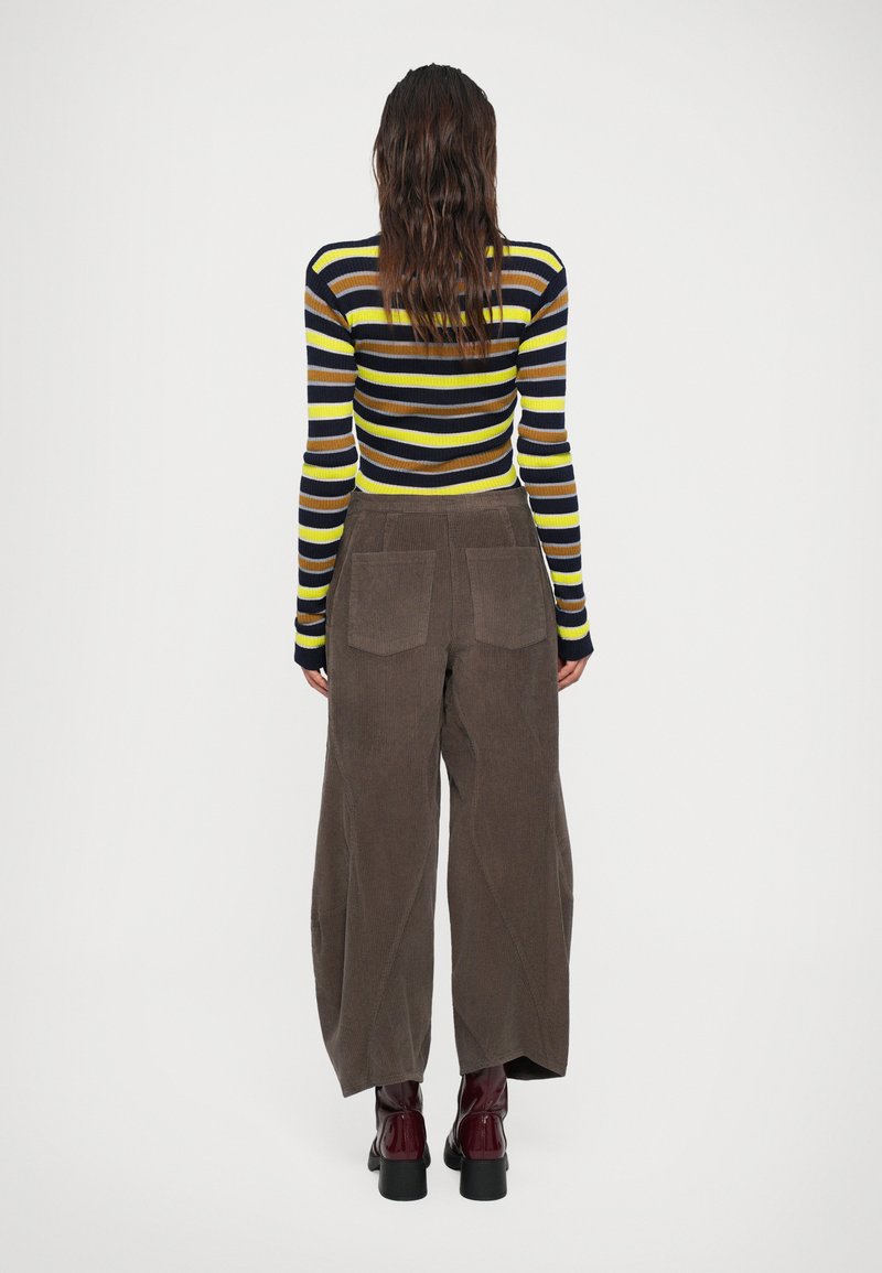 Donna con lunghi capelli castani, che indossa una maglietta a maniche lunghe a righe, pantaloni ampî in corduroy marroni e stivaletti con tacco bordeaux, di spalle.