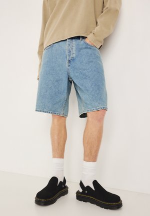 Hellblaue Jeansshorts mit lockerem Schnitt, ausgestattet mit einem klassischen Fünf-Taschen-Design und dezenten Verwaschungen. Kombiniert mit schwarzen Slippern.