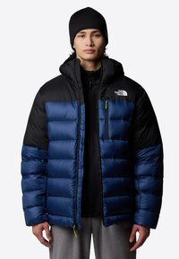 Niebieska i czarna kurtka puchowa z kapturem, z zamkniętą kieszenią na piersi, prążkowanymi mankietami oraz błyszczącym, teksturowanym wykończeniem. Logo marki na klatce piersiowej.
