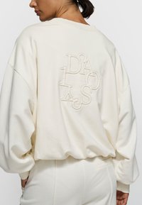 Sweat-shirt couleur crème avec un logo texturé en fil sur le dos. Comprend des poignets côtelés et une coupe décontractée.