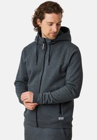 Grå zip-up hoodie med dragsko i huvan, mjuk textur, framfickor och ribbade ärmslut. Har en liten etikett nära fållen.