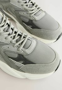 Baskets de sport grises avec un mélange de matériaux lisses et texturés, des panneaux en maille et des accents noirs avec une semelle blanche. Design à lacets.
