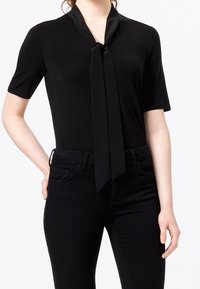 Musta, lyhythihainen paita, jossa on solmittava kaulustus, valmistettu sileästä kankaasta. Yhdistetty mustiin istuviin farkkuihin, luoden moniokromaattisen asun.