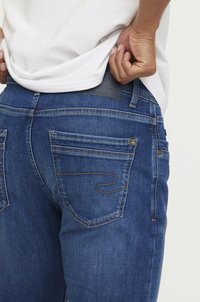 Jean en denim bleu foncé avec une texture lisse, cinq poches et des coutures contrastées. Présente une pièce en cuir à l'arrière de la taille.