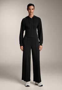 Fekete, rövidített kapucnis pulóver, elöl cipzárral és húzózsinórokkal, széles lábú fekete nadrággal párosítva. Sima textúra, minimális díszítés, laza dizájn.