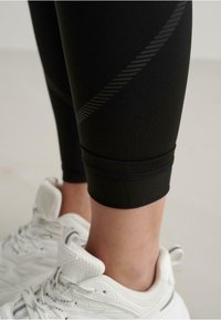 Colanți negri cu o textură netedă, având un model în dungi diagonale și o manșetă strânsă la gleznă, asortați cu pantofi sport albi.