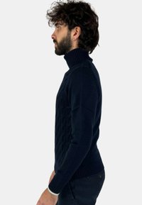 Maglione a collo alto lavorato a maglia di colore blu navy, caratterizzato da un motivo a diamante testurizzato e polsini e orlo a coste. Modello mostrato di lato.
