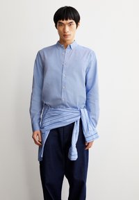 Light blue button-up shirt with a collar, paired with a striped blue shirt tied around the waist. Dark denim pants complete the outfit.