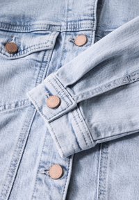 Giacca in denim blu chiaro con bottoni in bronzo, caratterizzata da cuciture visibili e un polsino ripiegato. La texture è leggermente usurata, con una finitura liscia.