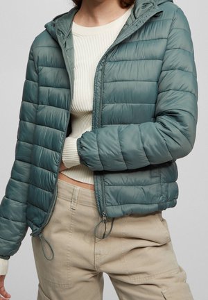 Person wearing a teal quilted jacket over a cream ribbed sweater and beige pants, showing midsection and zipped jacket partially open.