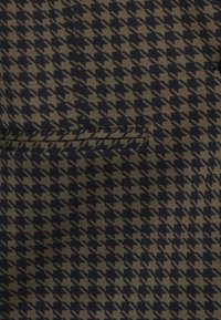 Närbild av brunt och svart hundtandsmönstrat tyg med en horisontell ficka, som visar den vävda texturen och mönsterdetaljer.