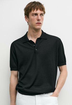 Junger Mann mit schwarzem kurzärmeligen Poloshirt und weißer Hose steht vor einem schlichten, hellen Hintergrund und blickt nach links.