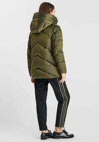 Olijfgroene pufferjas met capuchon, voorzien van chevronstiksel. Gecombineerd met zwarte gestreepte broeken en zwarte schoenen.