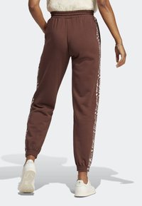 Person wearing brown joggers with white patterned side stripes and white sneakers, shown from the back against a white background.