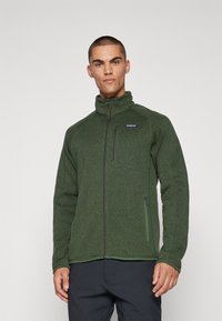 Dark green fleece jacket with high collar, full zip front, side pockets, ribbed cuffs, and a small logo on the chest. Soft texture overall.