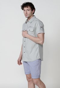 Camicia grigia chiaro a maniche corte con due tasche sul petto e dettagli in bottone, abbinata a pantaloni corti arrotolati viola chiaro, in piedi contro uno sfondo neutro.