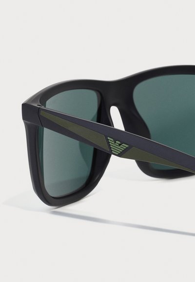 Ochelari de soare negri cu lentile de nuanță verde. Prezintă un design unghiular, o ramă neagră elegantă și un accent verde pe templu cu detalii ale logo-ului.