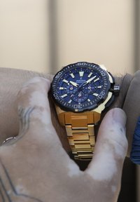 Montre chronographe noire et dorée avec un boîtier rond, un cadran bleu, des index lumineux et un bracelet à maillons dorés, portée au poignet.