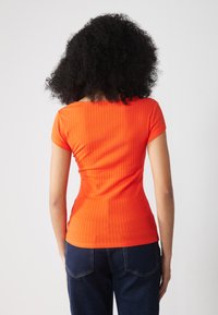 T-shirt realizzato in tessuto a coste, di colore arancione vivace, maniche corte, design aderente, indossato con jeans blu scuro. Dettaglio visibile sulla schiena.