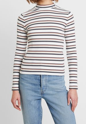 Femme portant un haut ajusté à manches longues blanc avec des rayures horizontales noires et rouges, associé à un jean bleu clair.