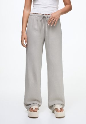 Femme portant un pantalon large gris clair avec taille à cordon, des sandales à plateforme blanches et un top court blanc.