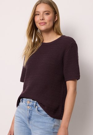 Jeune femme aux longs cheveux blonds portant un haut texturé violet foncé à manches courtes et un jean taille haute bleu clair.