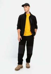 Sort fleecejakke med en lynlås lomme, gul t-shirt, sorte bukser med kontrasterende knælapper og brune sko. Sort kasket fuldender looket.