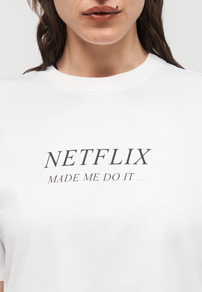 Γυναίκα που φοράει λευκό μπλουζάκι με το κείμενο "NETFLIX MADE ME DO IT" τυπωμένο με μαύρα γράμματα μπροστά, ορατό από τους ώμους ως το στήθος.