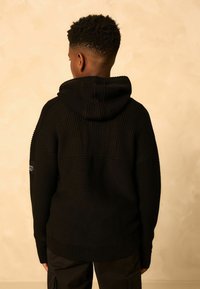 Haut à capuche en tricot noir avec un design en côtes texturé, doté d'une grande capuche attachée et de détails de couture minimalistes. Vue arrière présentée.
