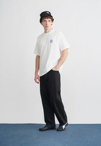 Biała bawełniana koszulka z małym fioletowym logo, zestawiona z luźnymi czarnymi spodniami i czarnymi butami z białym akcentem. Prosty, casualowy strój.