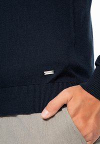Maglione blu navy a maglia con polsini e orlo a coste; presenta una piccola etichetta con logo argentato sul lato sinistro inferiore; texture liscia.