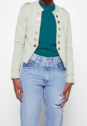 Persoon die een lichtgroene jas met gouden knopen, een blauwgroene blouse en lichtblauwe gescheurde spijkerbroek draagt, staande tegen een witte achtergrond.