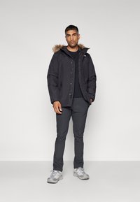 Zwarte geïsoleerde jas met een met bont afgewerkte capuchon, ritssluiting en knoopsluiting, gedragen over een zwart T-shirt, gecombineerd met donkergrijze broek en grijze sneakers.