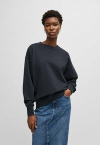 Navy sweatshirt with a relaxed fit, ribbed cuffs, and hem. Features a small embroidered logo on the chest. Paired with a denim skirt.