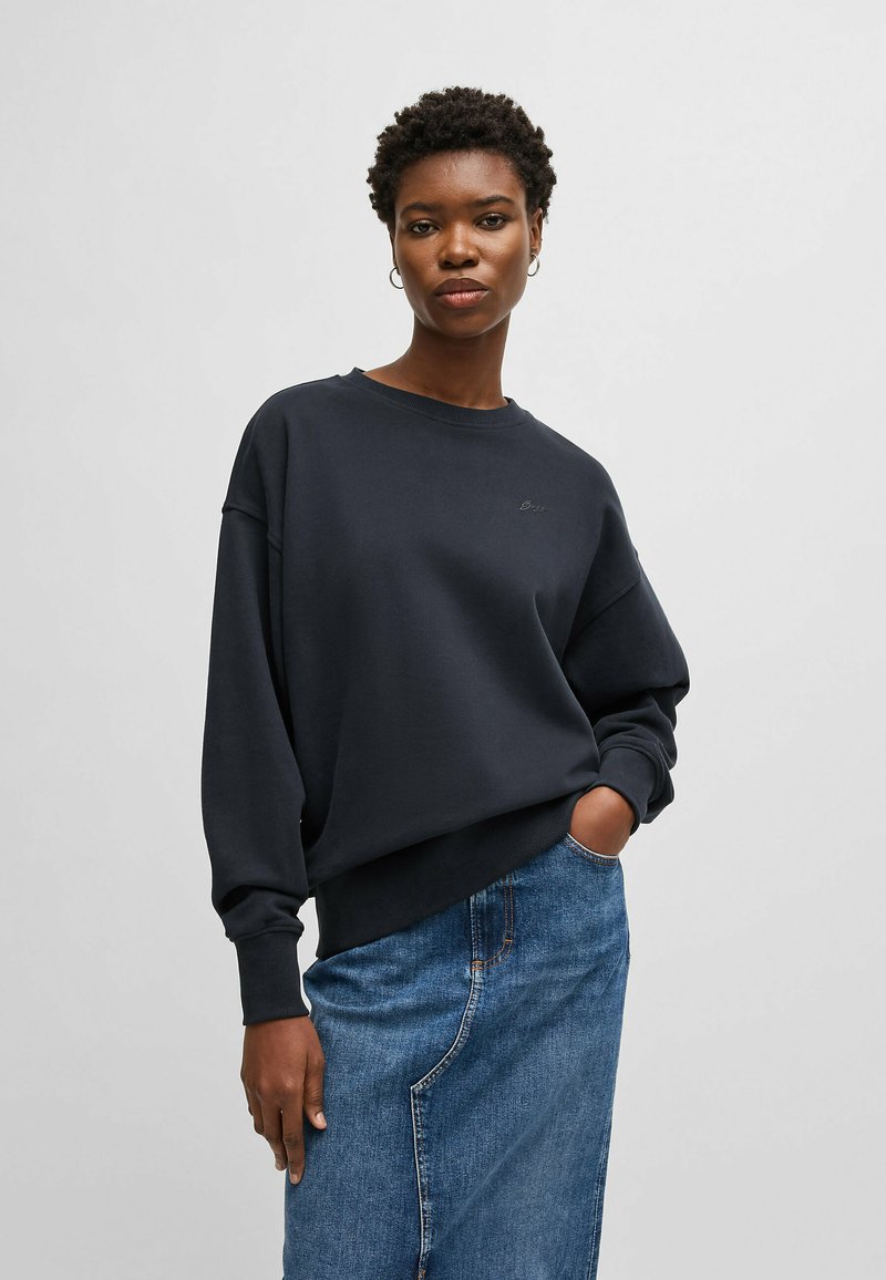 Navy sweatshirt with a relaxed fit, ribbed cuffs, and hem. Features a small embroidered logo on the chest. Paired with a denim skirt.