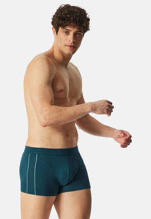 Man zonder shirt met krullend haar, gekleed in donkerteal boxershorts, zijwaarts staand tegen een effen lichte achtergrond.