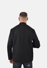 Giacca nera in misto lana con colletto a listino, maniche lunghe e design dritto sul retro, abbinata a jeans in denim, vista da dietro.