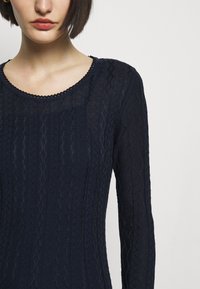 Námořnickě modré pletené tričko s dlouhým rukávem, které má texturovaný vzor, kulatý výstřih a složitou ozdobu podél výstřihu.