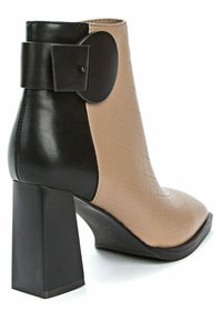 Botín con parte superior beige acolchada con detalles en negro, punta redondeada y tacón grueso de bloque. Detalle decorativo de moño negro en el costado.