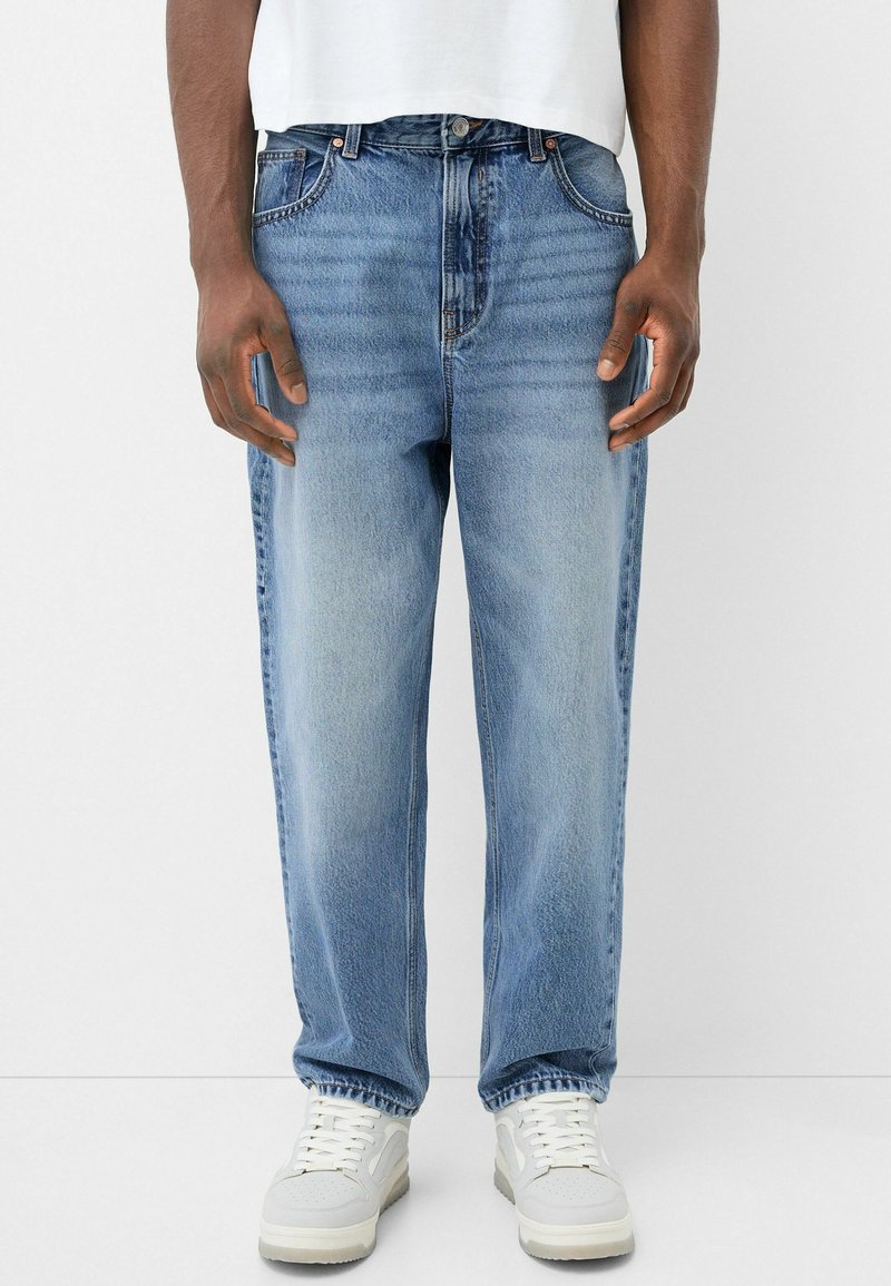 Jeans de mezclilla azul claro con un corte recto, diseño desgastado, cinco bolsillos y herrajes de metal, combinados con zapatillas blancas.