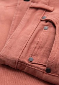 Rostfarbenes Gewebe mit strukturierter Oberfläche, versehen mit Metallknöpfen und detaillierten Nähten. Enthält ein Taschenklappen-Design.