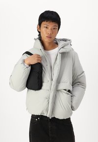 Chaqueta de plumas gris claro con capucha, bolsillos con cremallera y puños elásticos; combinada con un pequeño bolso negro cruzado y pantalones oscuros.