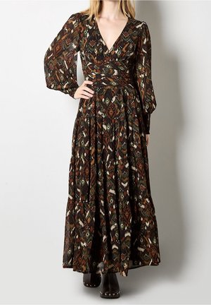 Kobieta w długiej sukience maxi z długim rękawem, dekoltem w serek i ciemnym plemiennym wzorem, z warstwową spódnicą i czarnymi, ćwiekowanymi botkami na prostym tle.