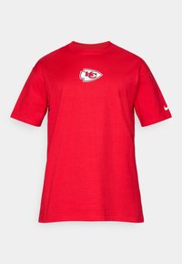 Sarkana kokvilnas T-kreklā ar apaļu kaklu, uz krūšu daļas ar baltu un pelēku Kansas City Chiefs logo un nelielu Nike swoosh uz piedurknes.